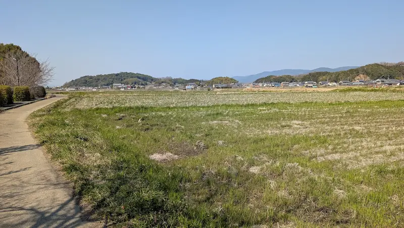 明日香村にある石神遺跡の全景。手前には当時の迎賓館の様子が描かれたカラーの案内板が立ち、その向こうには遮るもののない青々とした広い原っぱと、遠くの山々や民家が広がっている。
