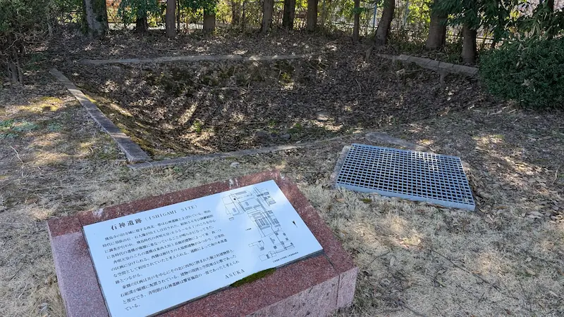 明日香村にある石神遺跡の解説看板。手前の御影石の台座に設置された看板には遺跡の概要や建物配置図が書かれており、奥には石組みで囲まれた四角い井戸跡の遺構とグレーチング(鉄格子)の蓋が見える。
