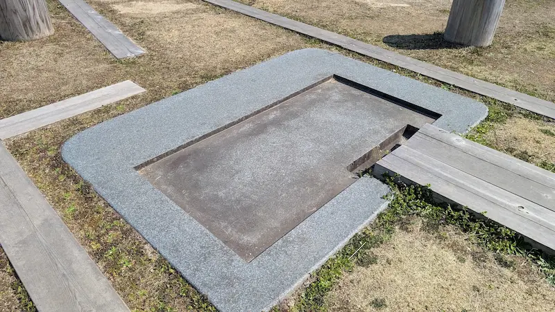 飛鳥水落遺跡の中心部にある、水時計の水槽が設置されていたとされる場所の復元遺構。グレーの御影石風の枠で囲まれた四角い窪みがあり、周囲には等間隔に配置された柱の土台や木製の通路が写っている。