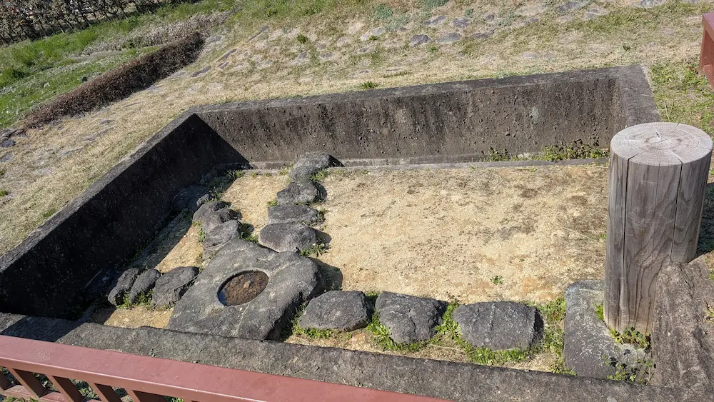 3月の明日香村にある飛鳥水落遺跡の様子。手前には遺跡の概要や水時計(漏刻)の仕組みが書かれた解説看板が立ち、奥には復元された四角いコンクリートと石組みの基壇が広がっている。
