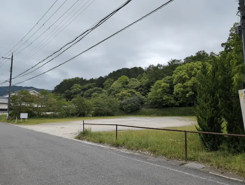 明日香村にある未舗装の広い有料駐車場。砂利敷きの平坦な土地で、区画線はない。背景には豊かな新緑の山々が広がり、手前には道路と仕切りのフェンスが見える。