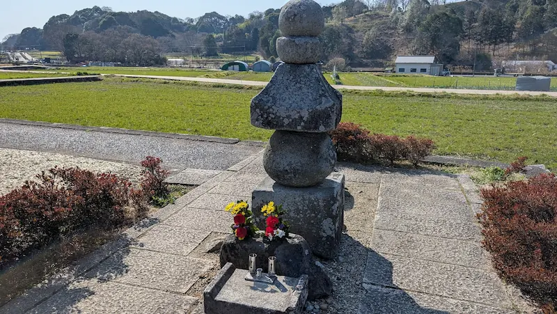明日香村の田園風景の中に立つ石造りの五輪塔。供えられた花が彩りを添え、周囲にはのどかな農地が広がっている様子。