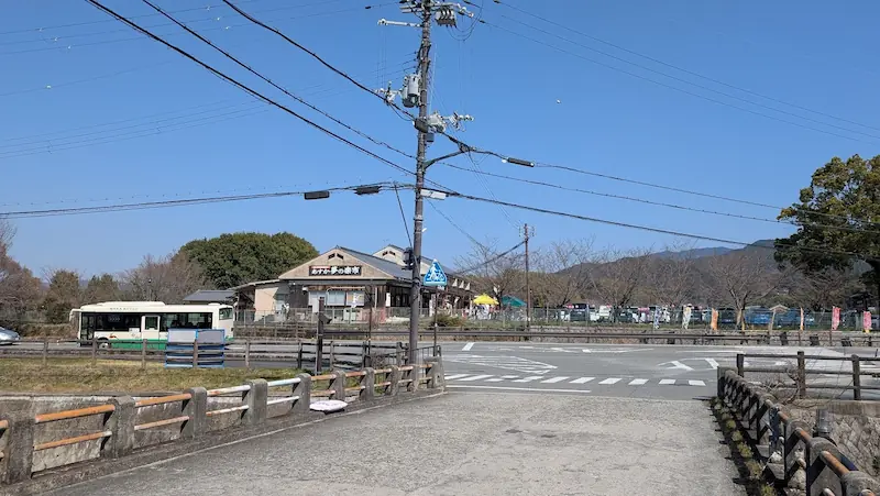 明日香村の橋の上から望む風景。手前には流れる飛鳥川と土手、その奥には木々に囲まれて「明日香夢の楽市」の建物が見え、背景には明日香村特有のなだらかな山並みが広がっている。