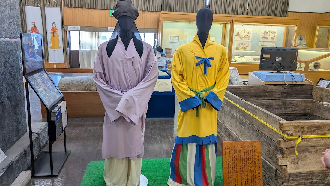 明日香村埋蔵文化財展示室の内部。飛鳥時代後半の衣装を身にまとった2体のマネキンが並んでおり、左は薄紫色の服に黒い帽子をかぶった男性役人の姿、右は黄色の上着にカラフルな縞模様のスカート（？の裳）を穿いた女性貴族の姿をしている。