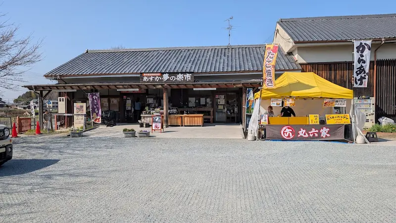 奈良県明日香村の直売所「あすか夢の楽市」の外観。広々とした砂利の駐車場と、たこ焼きや唐揚げの屋台が出店している店舗入り口の様子。