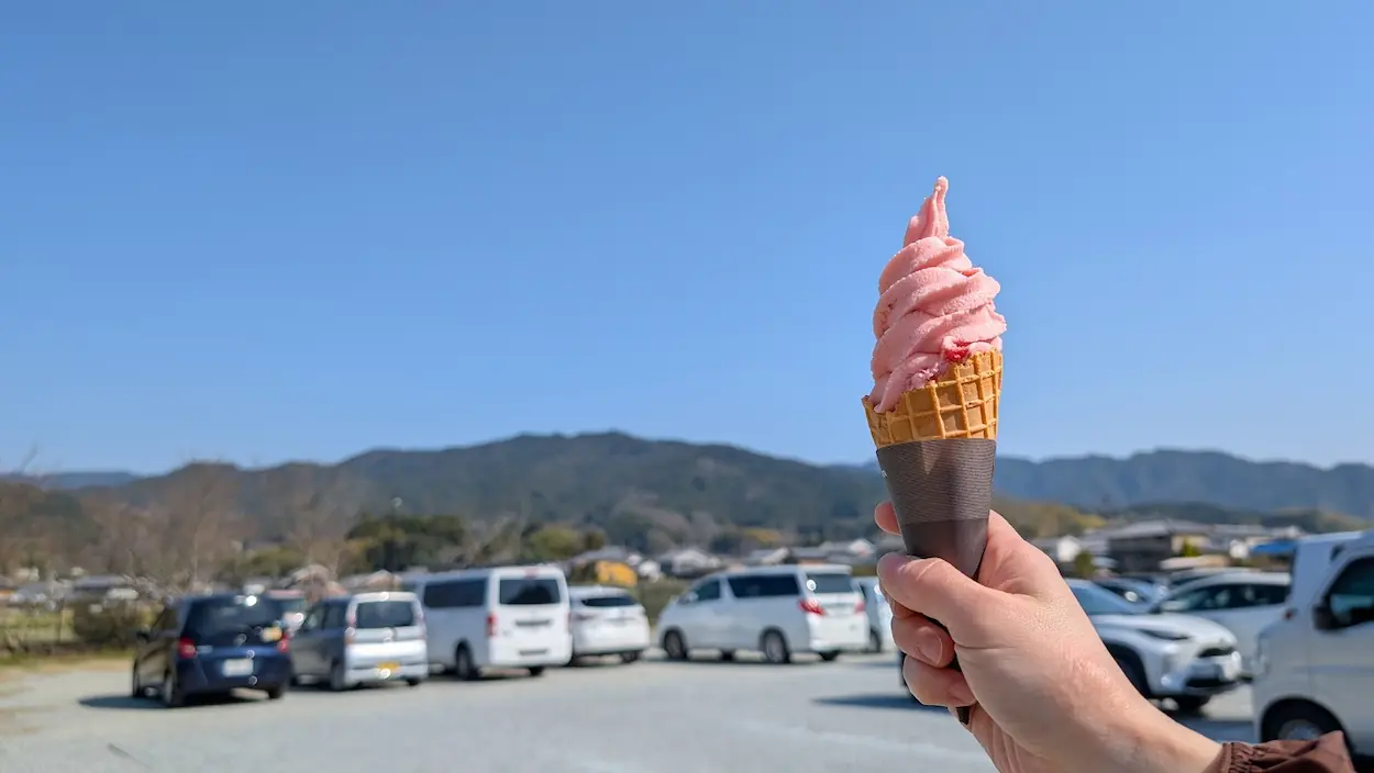 あすか夢の楽市の駐車場で、手に持ったあすかルビーのソフトクリーム。背景には青空と飛鳥の山々、そして満車の駐車場が広がり、春の観光シーズンの賑わいを感じさせる。