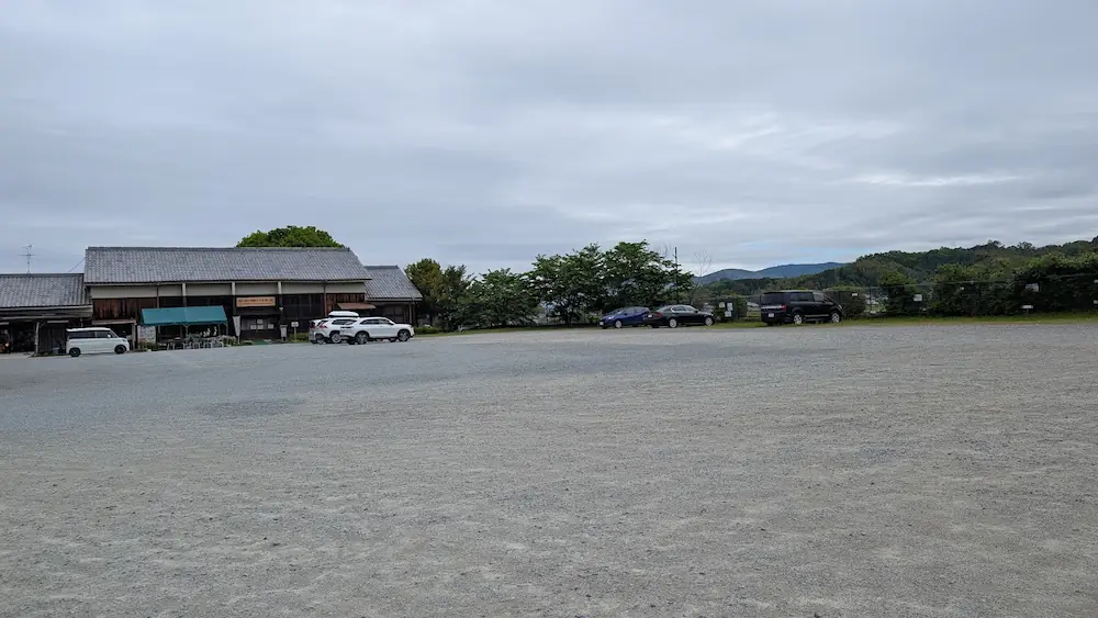 あすか夢の楽市の広大な無料駐車場。舗装された広いスペースがあり、周囲には新緑の山々と、あすか夢の楽市の建物が見える。