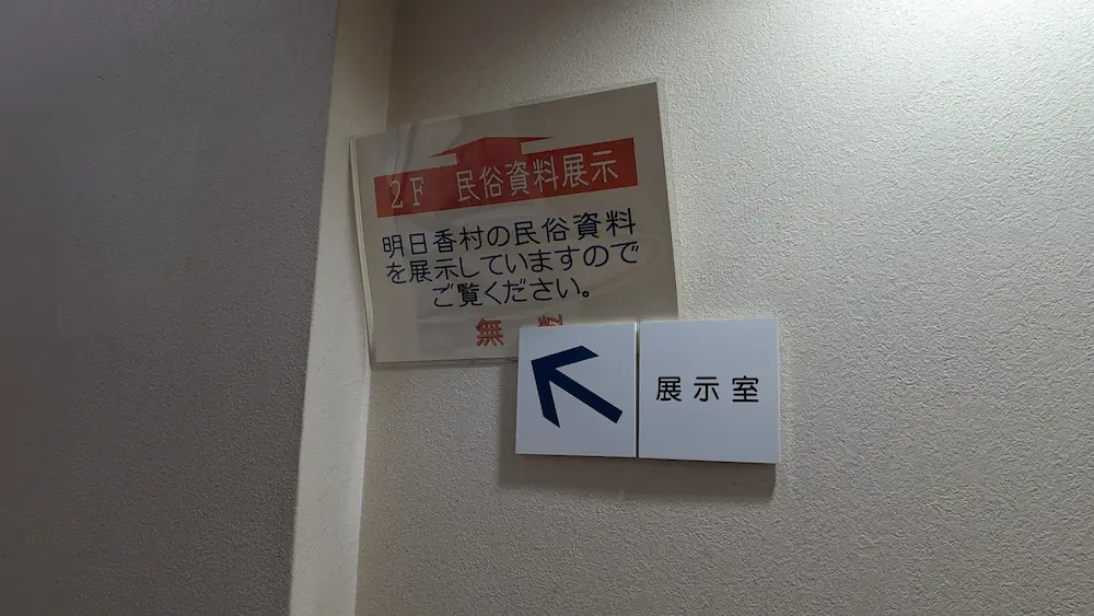 あすかむ館内の壁に貼られた案内。「2F 民俗資料展示」「明日香村の民俗資料を展示していますのでご覧ください」という文字と、赤字で「無料」と書かれた貼り紙、その下に左上を指す矢印と「展示室」の看板。