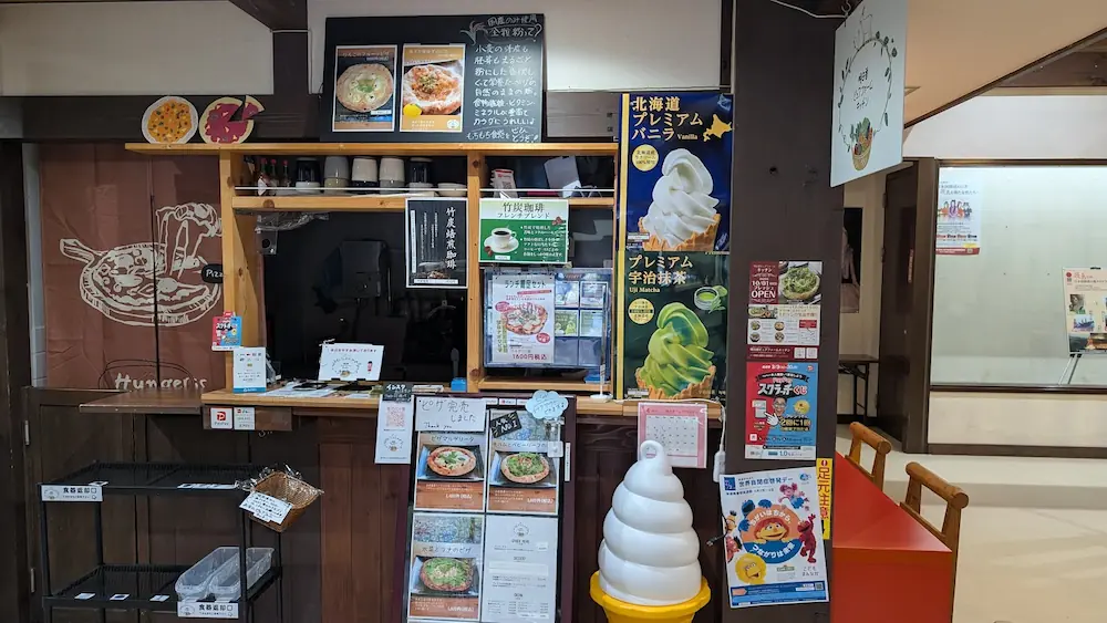 ASUCOME（あすかむ）内にある「明日香ビオファームキッチン」の注文カウンター。全粒粉ピザや竹炭焙煎珈琲のメニューが掲示されているが、中央には「ピザ完売しました」と書かれた貼り紙が出されている。