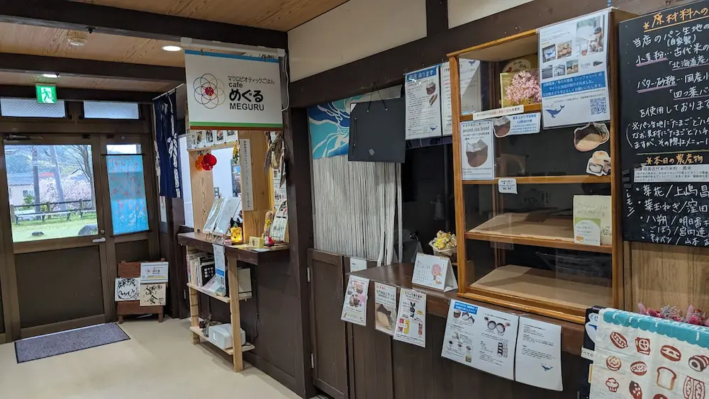 ASUCOME（あすかむ）内にある「cafe めぐる MEGURU」の店舗外観。木目を基調としたカウンターに看板が下がり、右側には原材料のこだわりが書かれた黒板と空のショーケースが並んでいる。左奥のガラス扉からは外の景色が見える。