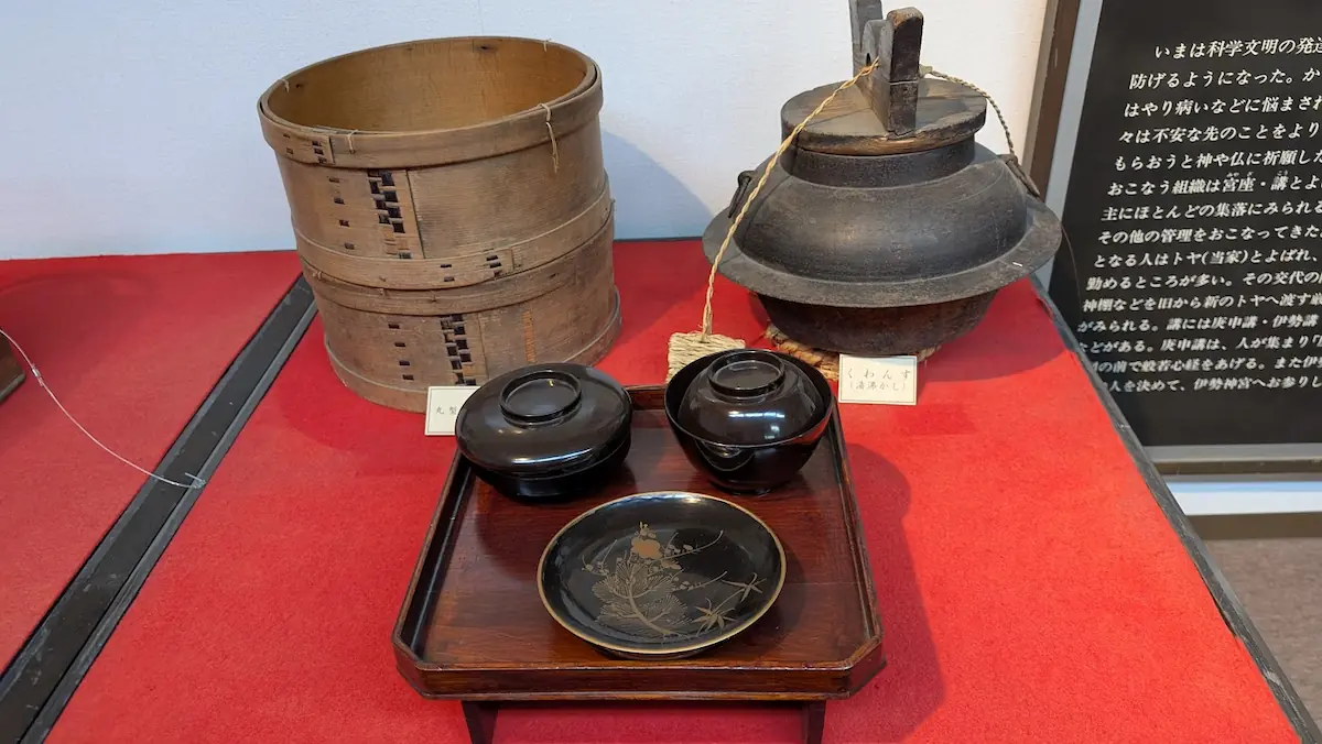 明日香民俗資料館の展示。赤い布の上に、木製のおひつ、鉄製の湯沸かし茶釜、漆塗りのお膳と吸物椀が並べられている様子。