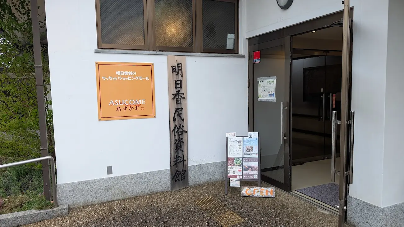 明日香村にある「明日香民俗資料館」とショッピングモール「ASUCOME（あすかむ）」の共通入り口。白い壁に木製の看板とオレンジ色の看板が並び、足元には立て看板が置かれている。