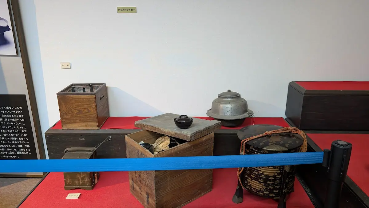 明日香民俗資料館の展示。赤い布の上に、大きな木箱や漆塗りの円筒形の容器、金属製の釜などが並んでいる。手前には青い進入禁止のベルトが張られている。