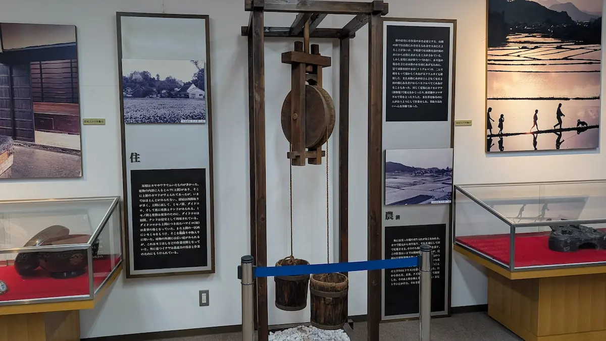 明日香民俗資料館の展示エリア。中央に木製の井戸滑車と桶が再現され、左右の壁には「住」と「農」に関する解説パネルと当時の生活風景写真が並んでいる。