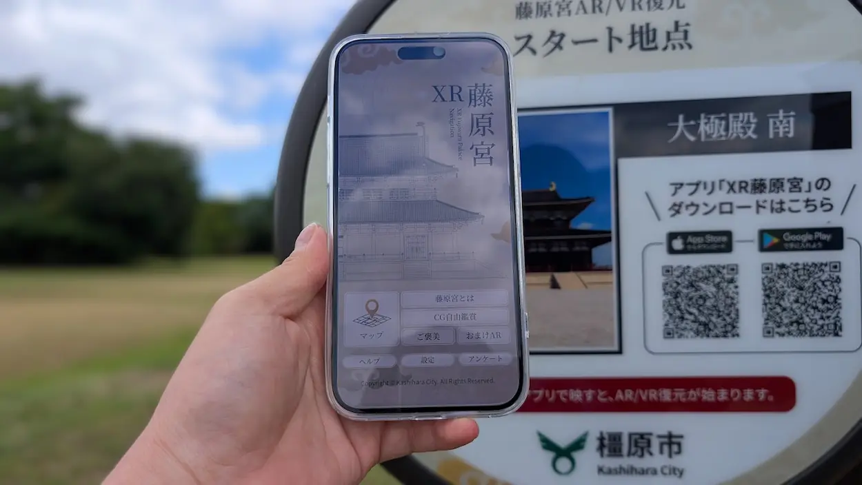 藤原宮跡のAR/VR復元スタート地点を示す看板の前で、スマホに「XR藤原宮」アプリのホーム画面を表示している様子。