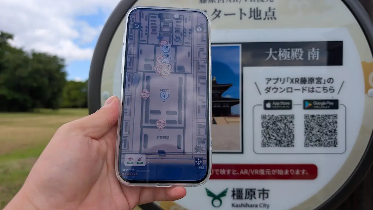 藤原宮跡にて、スマホで「XR藤原宮」アプリのマップ画面を表示し、アプリ起動用QRコードが記載された標識にかざしている様子。