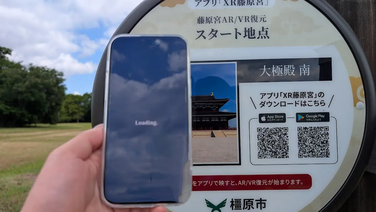藤原宮跡のスタート地点にある大極殿の写真マーカーにスマホをかざし、「XR藤原宮」アプリがARデータを読み込んでいる様子。