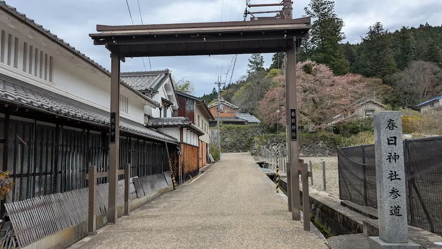 奈良県宇陀市にある春日神社の参道入り口。石造りの社標と木製の門が立ち、奥には城下町の家並みと山々の緑が広がっている。