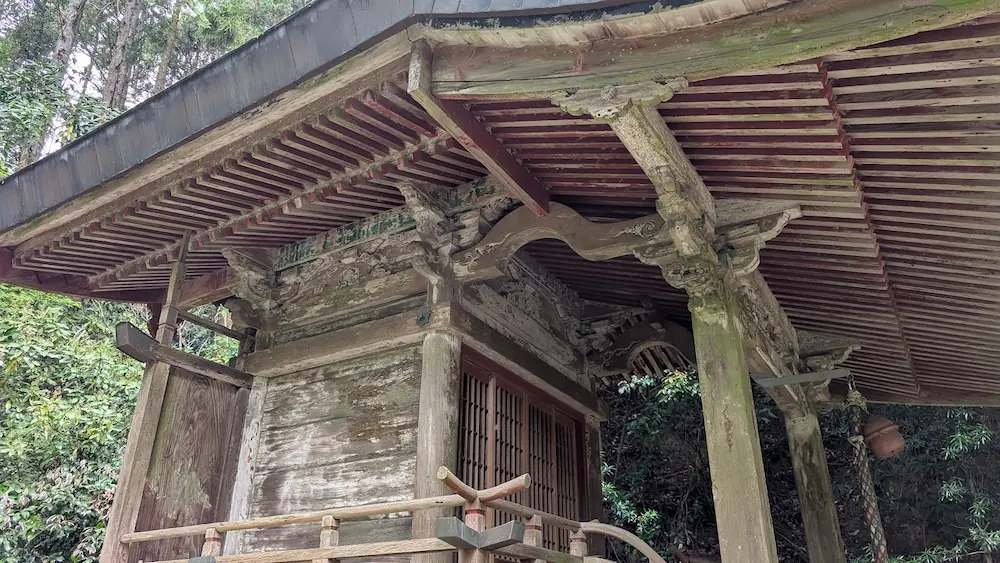 葛城水分神社本殿の木造建築のクローズアップ。年月を経て色が剥げた彫刻、複雑に組まれた軒下の垂木、緑の苔がうっすらと付いた柱の様子。