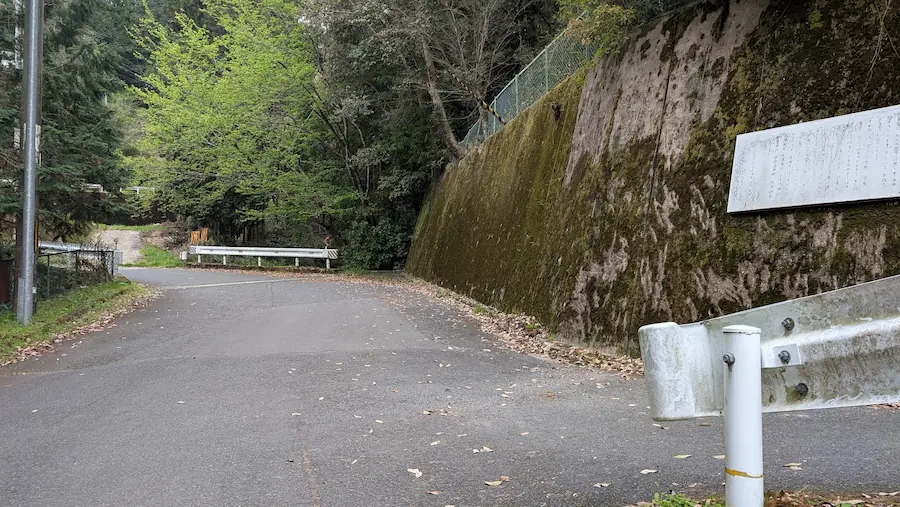 葛城水分神社へと続く細い舗装路と、右側の苔むした擁壁に設置された看板、手前のガードレールの様子。