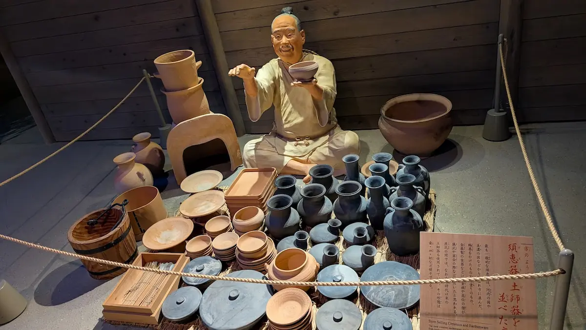 万葉文化館の展示。古代の市場で、灰色をした須恵器と赤茶色の土師器を大量に並べ、客に器を差し出す商人の人形ジオラマ。