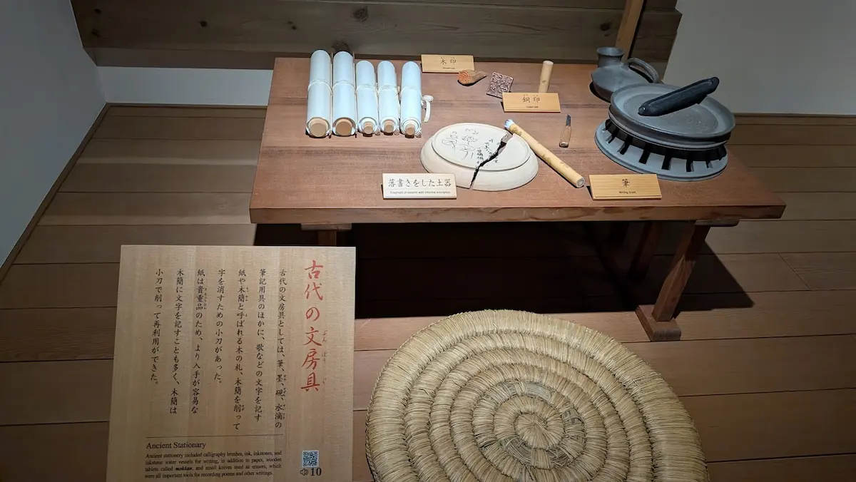 万葉文化館の展示。木製の机の上に、巻物、筆、硯、墨、そして人物の顔が描かれた「落書きをした土器」が並んでいる。手前には円形の円座（わろうだ）がある。