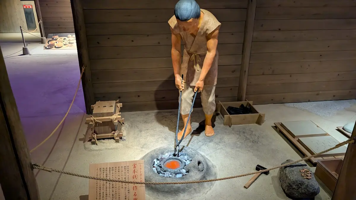 万葉文化館の展示。富本銭を作る職人の実物大ジオラマ。火床（ひどこ）で赤く溶けた銅を扱い、周囲には鋳型（いがた）や道具が置かれている様子。