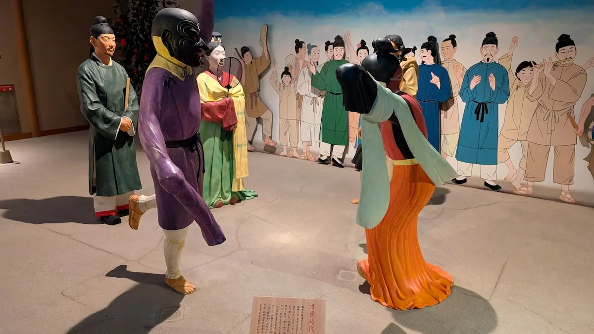 万葉文化館の展示。万葉時代の芸能を再現した実物大ジオラマで、特徴的な仮面と紫の衣装を纏った踊り手と、それを見守る貴族や観客（イラスト）の様子。