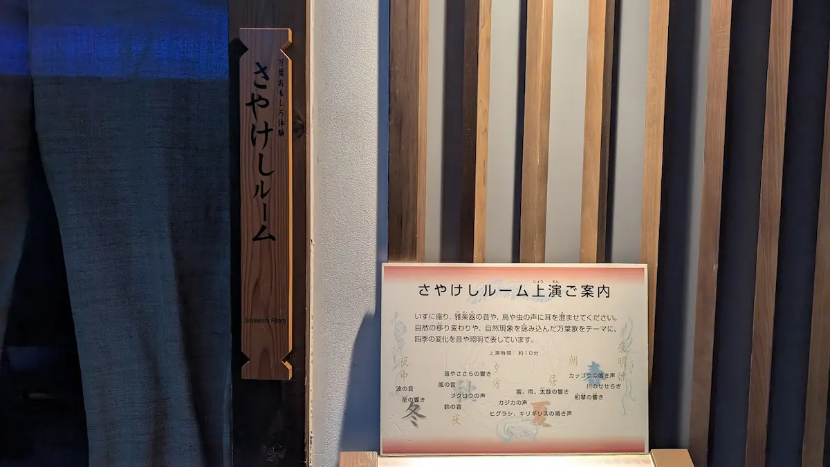 奈良県立万葉文化館内の「さやけしルーム」入り口。木製の看板と、上演内容（四季の音と照明の演出）を説明するパネルが設置されている様子。