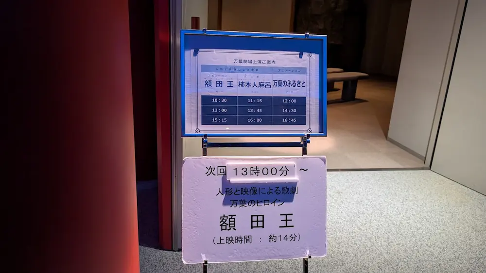 万葉劇場の「万葉劇場上演ご案内」看板。額田王、柿本人麻呂、アニメーションの上演時間が一覧表になっており、次回上演が「13時00分 額田王」であることを示している。