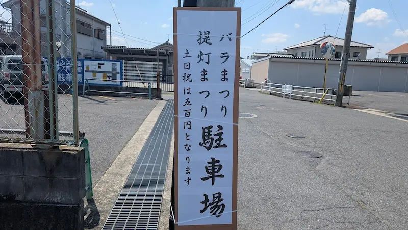 お房観音の駐車場入り口に立つ立て看板。「バラまつり 提灯まつり 駐車場 土日祝は五百円となります」と手書きの文字で案内されている。
