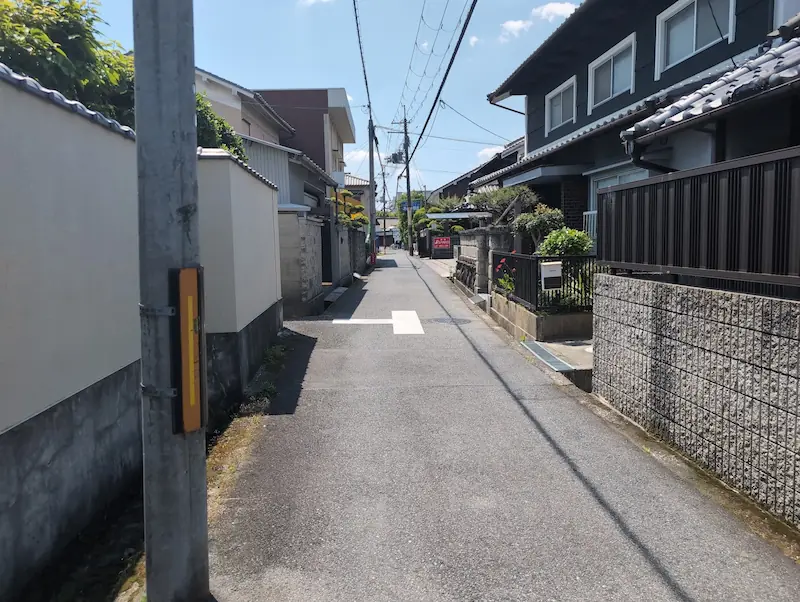 お房観音へと続く非常に狭い住宅街の道路。車1台が通るのがやっとの道幅で、両側には住宅の塀や壁が迫っており、対向車とのすれ違いが不可能な様子が写っている。