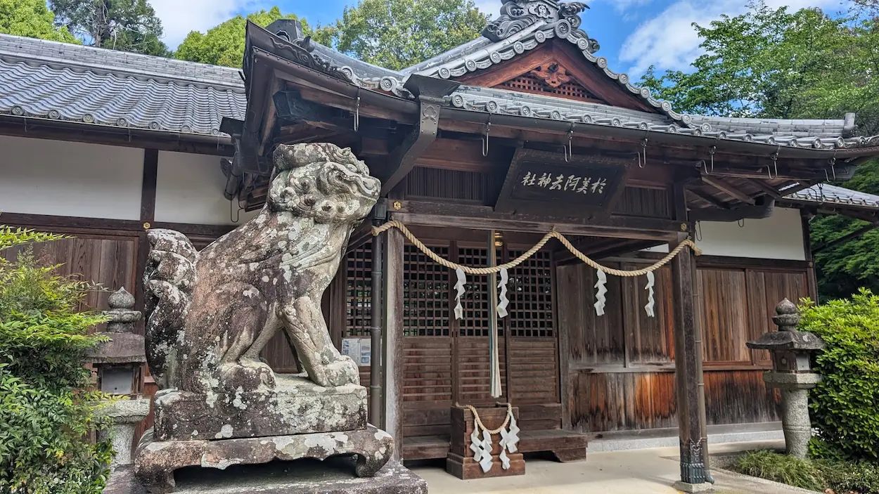 於美阿志神社の境内で、古風な石造りの狛犬と重厚な木造の本殿を至近距離から撮影した様子。
