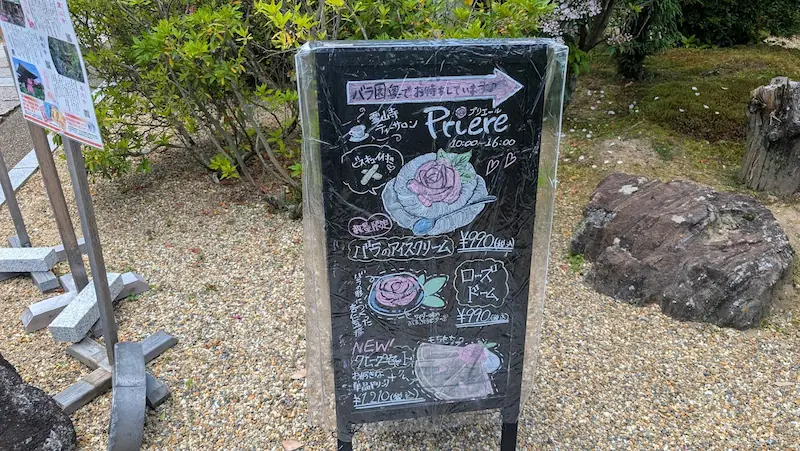 霊山寺バラ園の奥にあるティーラウンジ「プリエール」の入り口看板。白いイーゼルにバラの花が添えられ、ケーキセットやバラのアイスクリームなどのメニューが案内されている。