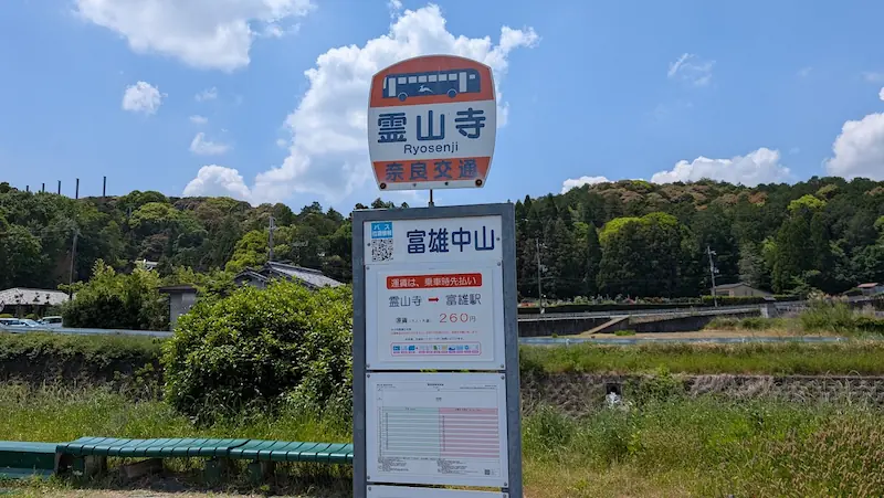 霊山寺（富雄中山）バス停の看板と時刻表。近鉄富雄駅行きバスの運賃が260円であることや、運行本数が1時間に1本程度と非常に少ないことが確認できる。背景には5月の青空と緑の山。