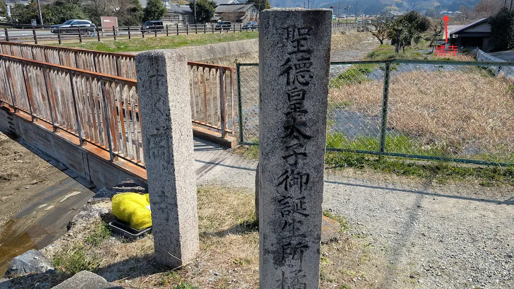 明日香村にある橘寺「聖徳皇太子御誕生所」と刻まれた歴史ある石碑。石碑の周りには苔むした石や美しい植栽が配置され、遥か奥には橘寺が。矢印とテキストで編集し位置を示している。