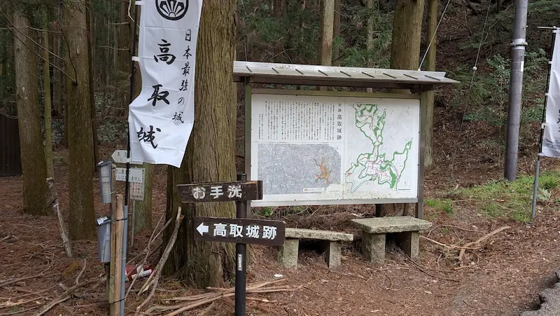 高取城跡の登城口にある大きな案内板。城の歴史と縄張図(マップ)が掲載されており、手前には「高取城跡」「お手洗」を指す木製の矢印看板と、白い幟が立っている。案内板の下には石造りの小さなベンチがある。