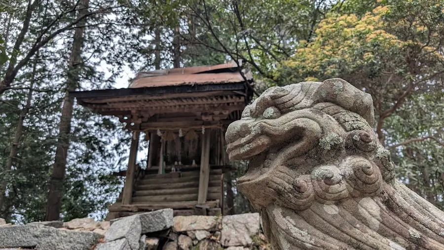 高取城跡の境内にある八幡神社。手前に大きく写った狛犬が、奥の石垣の上に建つ木造の本殿(トタン屋根)を滑稽な表情で見上げているような構図。