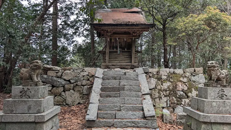 高取城跡の境内にある八幡神社の全景。石垣の上に木造の小さな社殿が建ち、手前には急な石段と一対の狛犬が配置されている。周囲は深い森に囲まれ、ひっそりとした佇まい。
