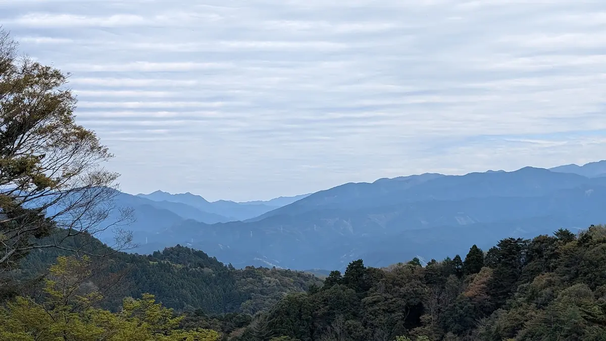高取城跡の本丸跡から望む山々のパノラマ風景。手前には深く茂る原生林の緑があり、遠景には青く霞んだ大峯連山の稜線が幾重にも重なって広がっている。