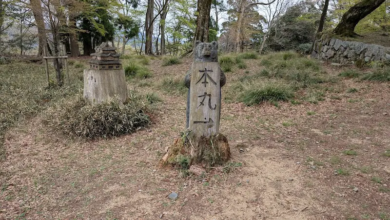 高取城跡の本丸前にある、切り株を加工した木彫りのオブジェ。左側には高取城の天守閣を模した模型、右側には「本丸」という文字を抱えた熊の彫刻が立っている。背後には石垣の一部が見える。