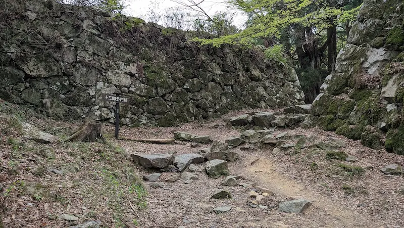 高取城跡の十三間多門跡。道の両側に高く積まれた野面積みの石垣がそびえ、中央には「十三間多門跡」の標識が立っている。地面は土と不揃いな石が露出し、歴史の重みを感じさせる遺構の風景。