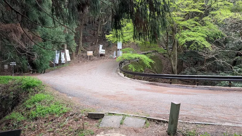 高取城跡の登城口付近にある未舗装の駐車スペース。道が大きくカーブし、路肩が広くなっている場所に車を停める形。周辺には案内看板や幟が立ち、木々に囲まれている。ここから先は車両進入禁止の標識がある。