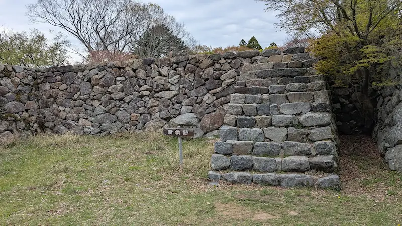 高取城跡の新櫓跡。野面積みの石垣が壁のように続き、右側には石を積み上げた急な階段(雁木)がある。手前には「新櫓跡」の小さな木製看板が立ち、地面は芝生と土が混ざっている。