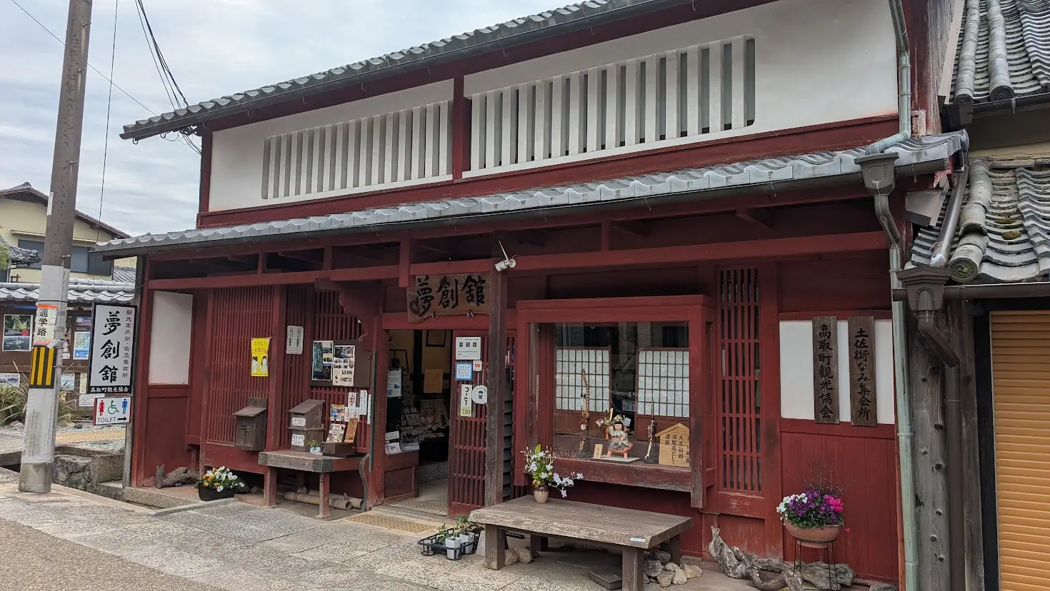 奈良県高取町の土佐街道沿いにある観光案内所「夢創舘」の外観。赤茶色の格子と白い漆喰壁が特徴的な伝統的な町家造りの建物で、入り口には木製の看板や観光案内、休憩用のベンチが置かれている。