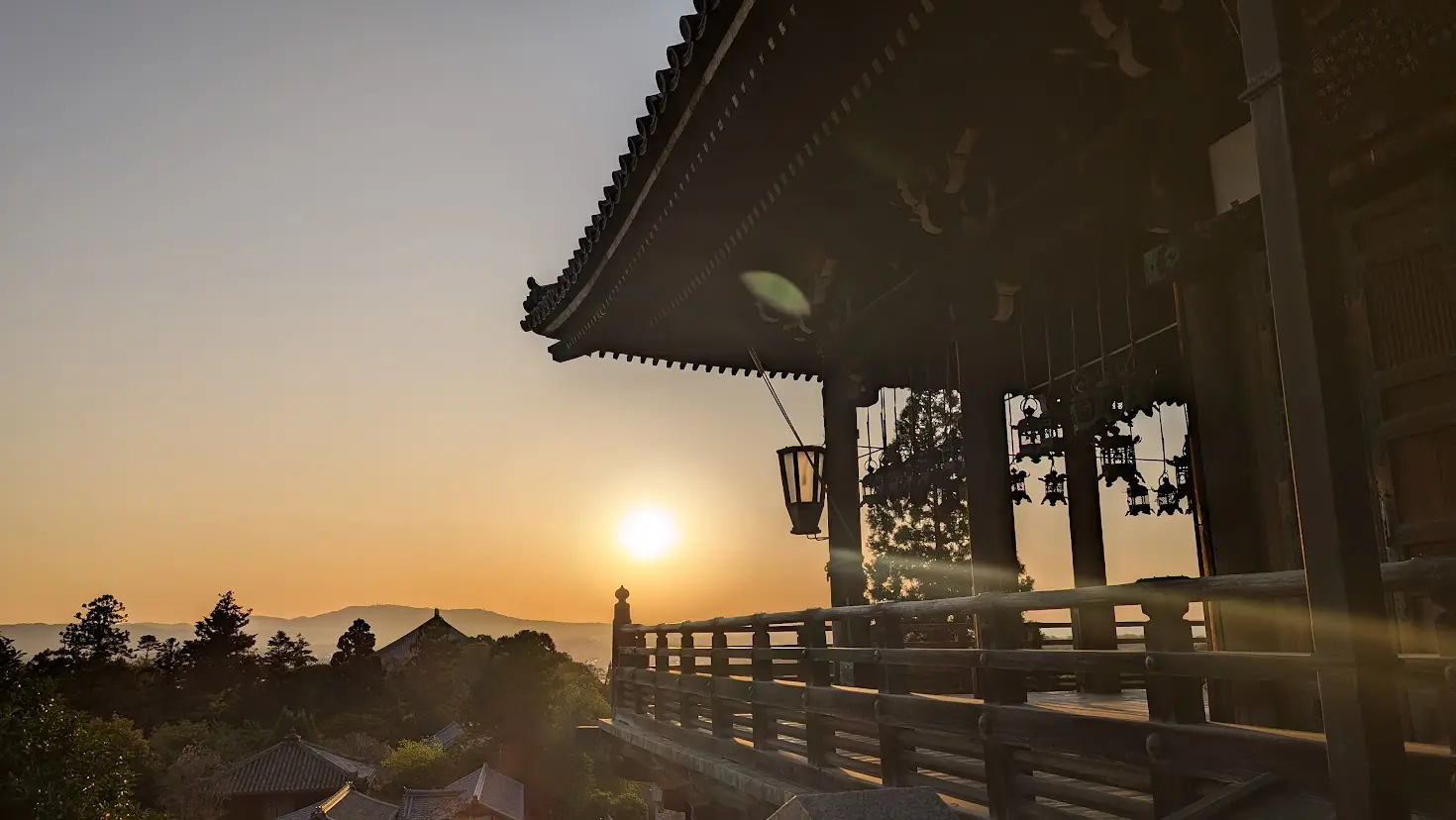 東大寺二月堂の舞台に吊るされた銅製の釣り灯籠。夕陽の光を浴びて細かな細工が黄金色に輝き、背後には舞台の木造建築が静かに佇んでいる
