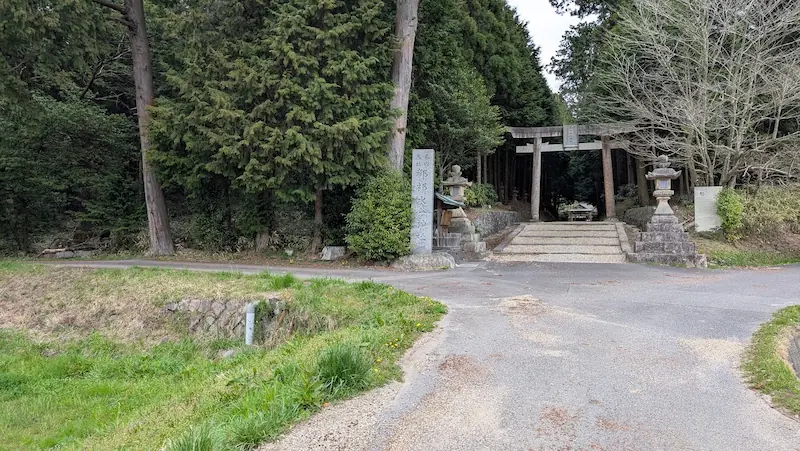 都祁水分神社の入り口を遠目から見た全景。道が二手に分かれる角に石造りの社標と鳥居が立ち、その後ろには鬱蒼とした高い杉木立が広がっている風景
