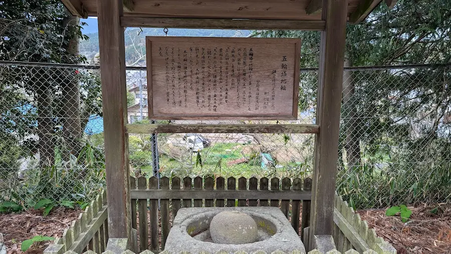 春日神社の境内にある、鎌倉時代の「五輪塔地輪(ごりんとうちりん)」。木製の屋根付き案内板の下に、丸い石が置かれた四角い石造物が祀られている。背景には金網越しに街並みが広がっている。