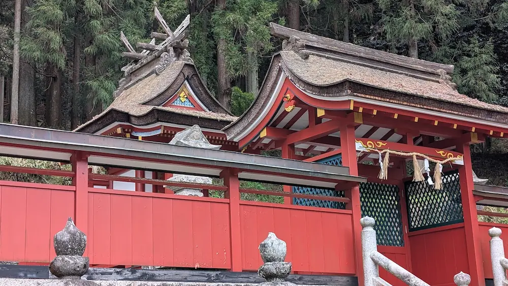 奈良県宇陀市・春日神社の本殿。美しい曲線を描く茅葺屋根と、朱色に彩られた柱や壁が特徴的。背後には深い杉林が広がり、神聖な雰囲気を醸し出している。