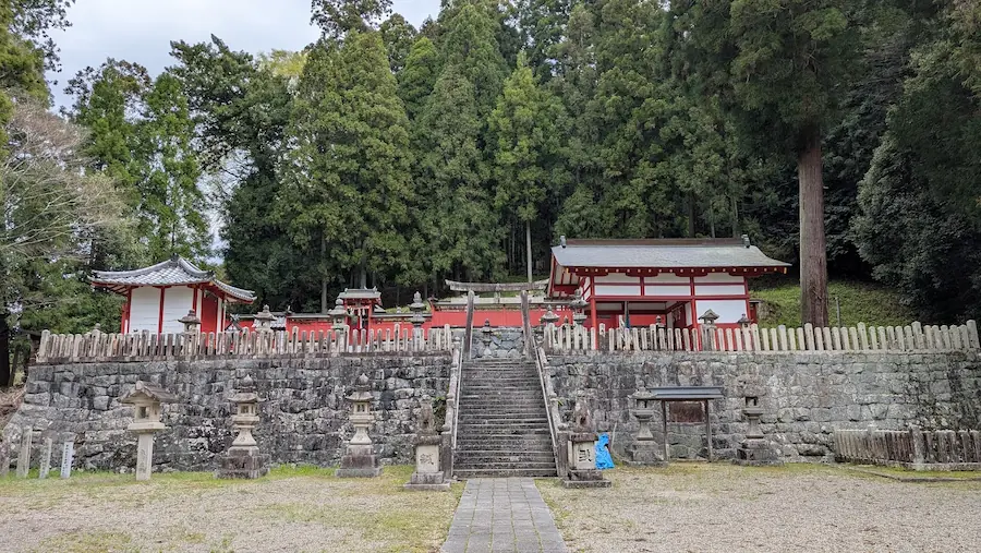 奈良県宇陀市にある春日神社の全景。高い石垣の上に朱塗りの社殿が建ち、手前には石灯籠が並んでいる。背後には鬱蒼とした森が広がっている。